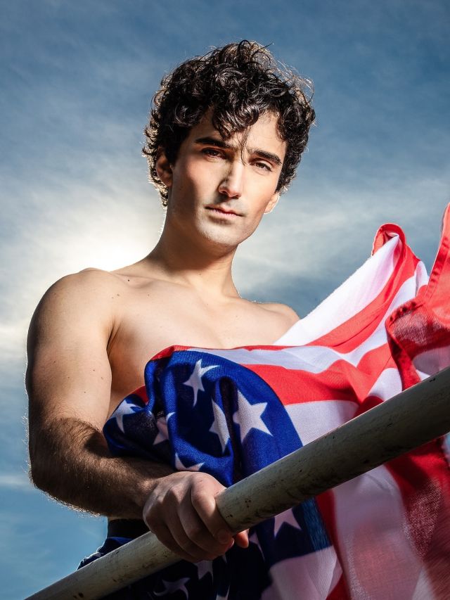 AMERIKA I Emre Aksızoğlu fotografiert von Esra Rotthoff; Im Zentrum des Fotos steht ein junger Mann (Schauspieler), um die 30 Jahre, mit blassem Teint und dunklen Locken. Mit leicht geneigtem Kopf blickt er in die Kamera, die ihn leicht von unten, ab Hüfthöhe fotografiert. Sein nackter Oberkörper ist von der Brust abwärts durch eine im Wind wehende Amerika-Fahne verdeckt. Die rechte Hand umgreift ein Geländer. Sein Blick deutet direkt in die Kamera. Im Hintergrund ist ein sonniger Himmel.